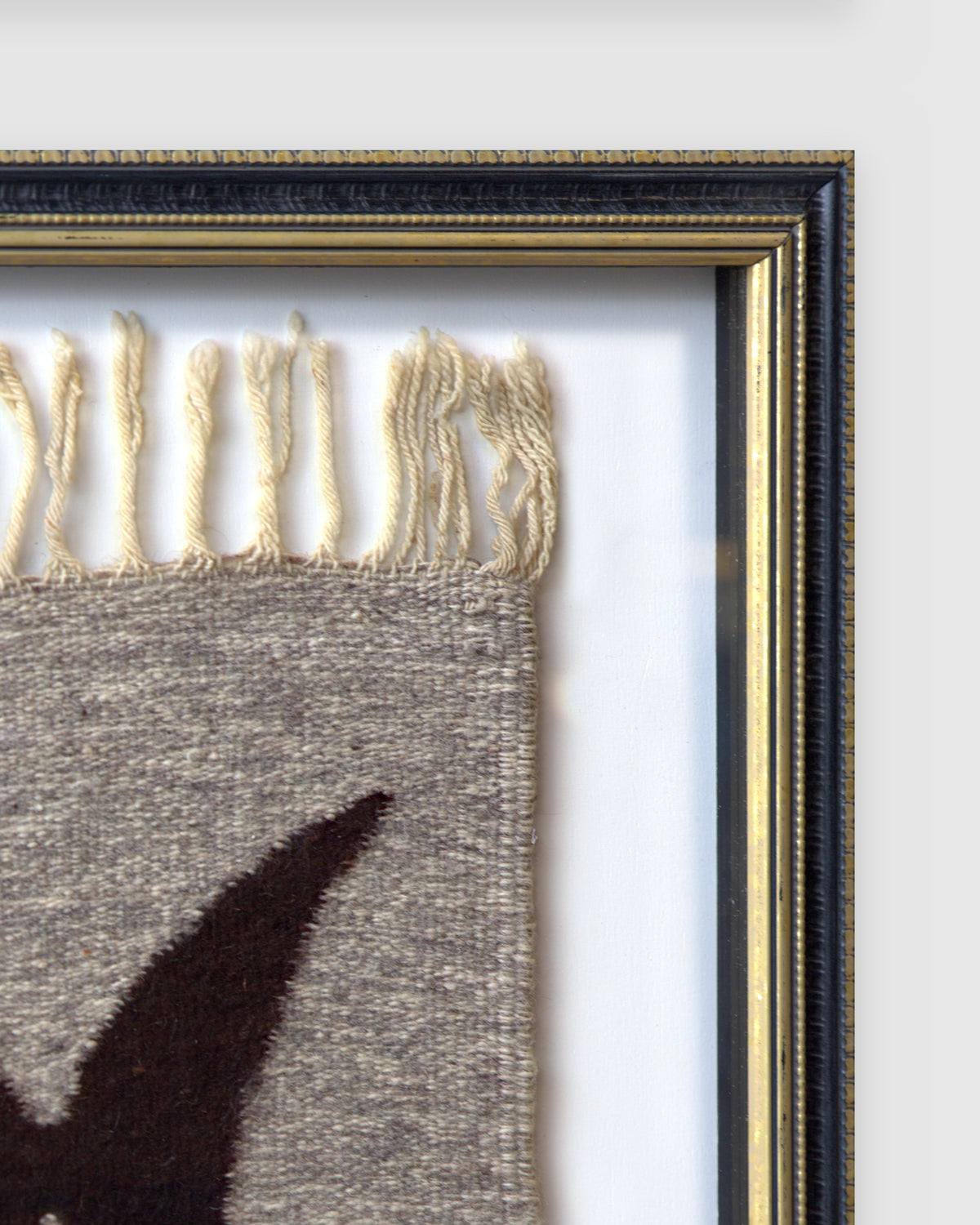 Upper right corner of the black and gold frame, with the tapestry fringe and white mat behind glass, on a light gray background.