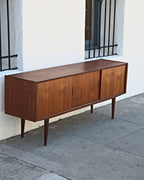 Three-quarter view of a mid century Danish teak credenza with three sliding doors and tapered legs against a white stucco wall
