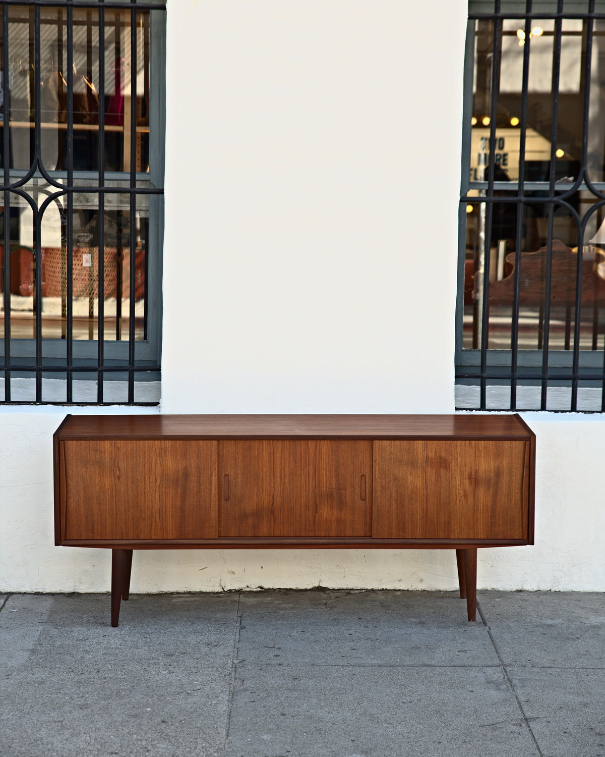 Front view of a long, low Danish teak credenza between two barred shop windows