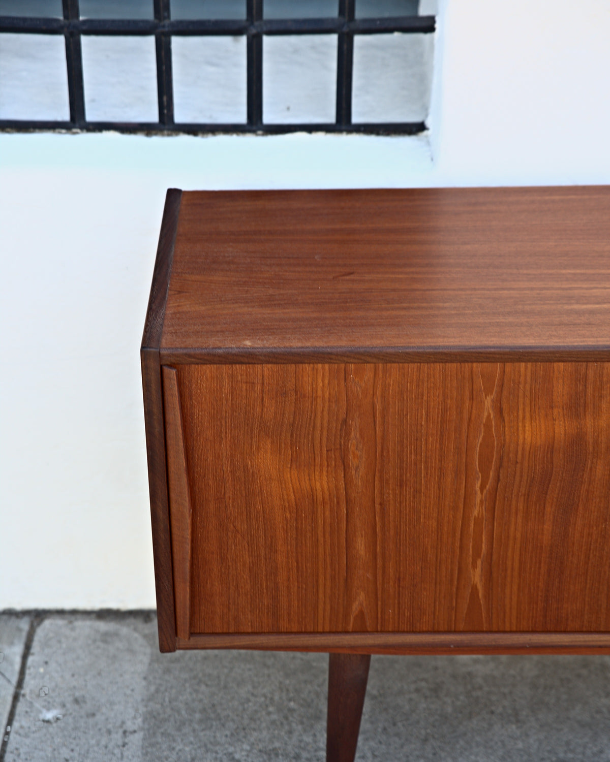 Corner of a teak credenza showing the edge-banded top, sliding door and tapered leg