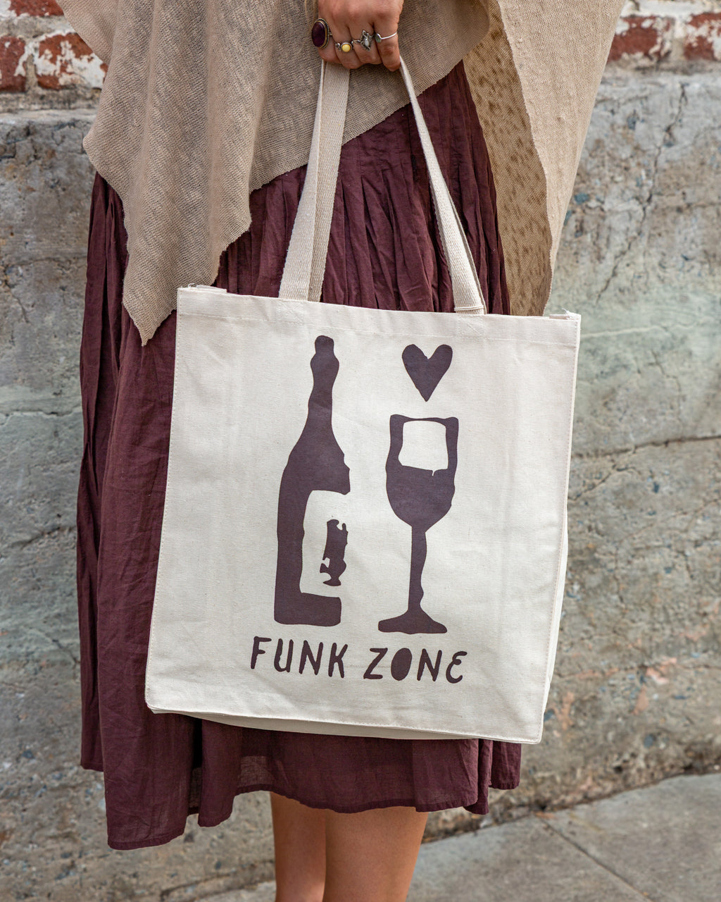 Natural canvas Funk Zone tote bag screen printed in burgundy with a wine bottle, glass and heart, carried at the side against a concrete wall.