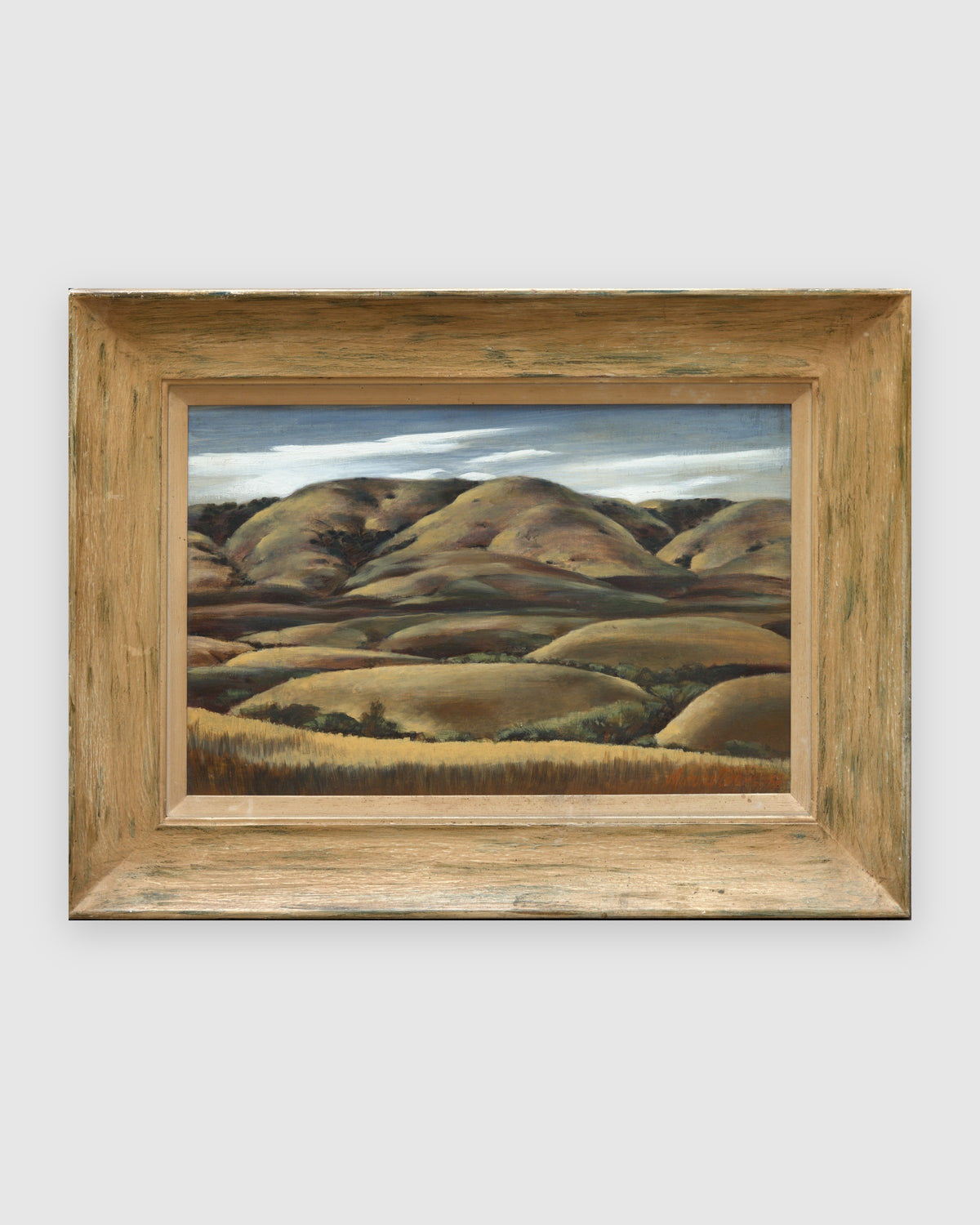Straight-on view of a Milford Zornes painting Hills of Nipomo, golden rolling hills under gray clouds, in a wide limed wood frame, on a light gray background.